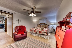 Une chambre à coucher avec un lit, une chaise rouge, un ventilateur de plafond, une commode en bois et une croix accrochée au mur.