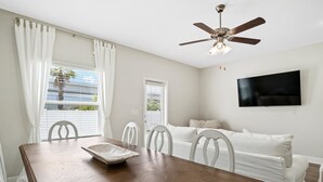A dining area with a wooden table, white chairs, a ceiling fan, and a TV mounted on the wall.