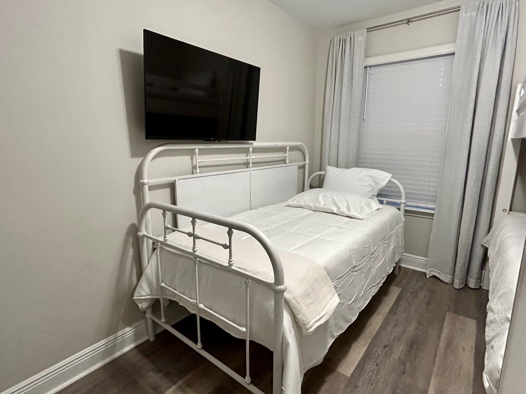 A single-bed room with a television mounted on the wall, a window with curtains, and a wooden floor.