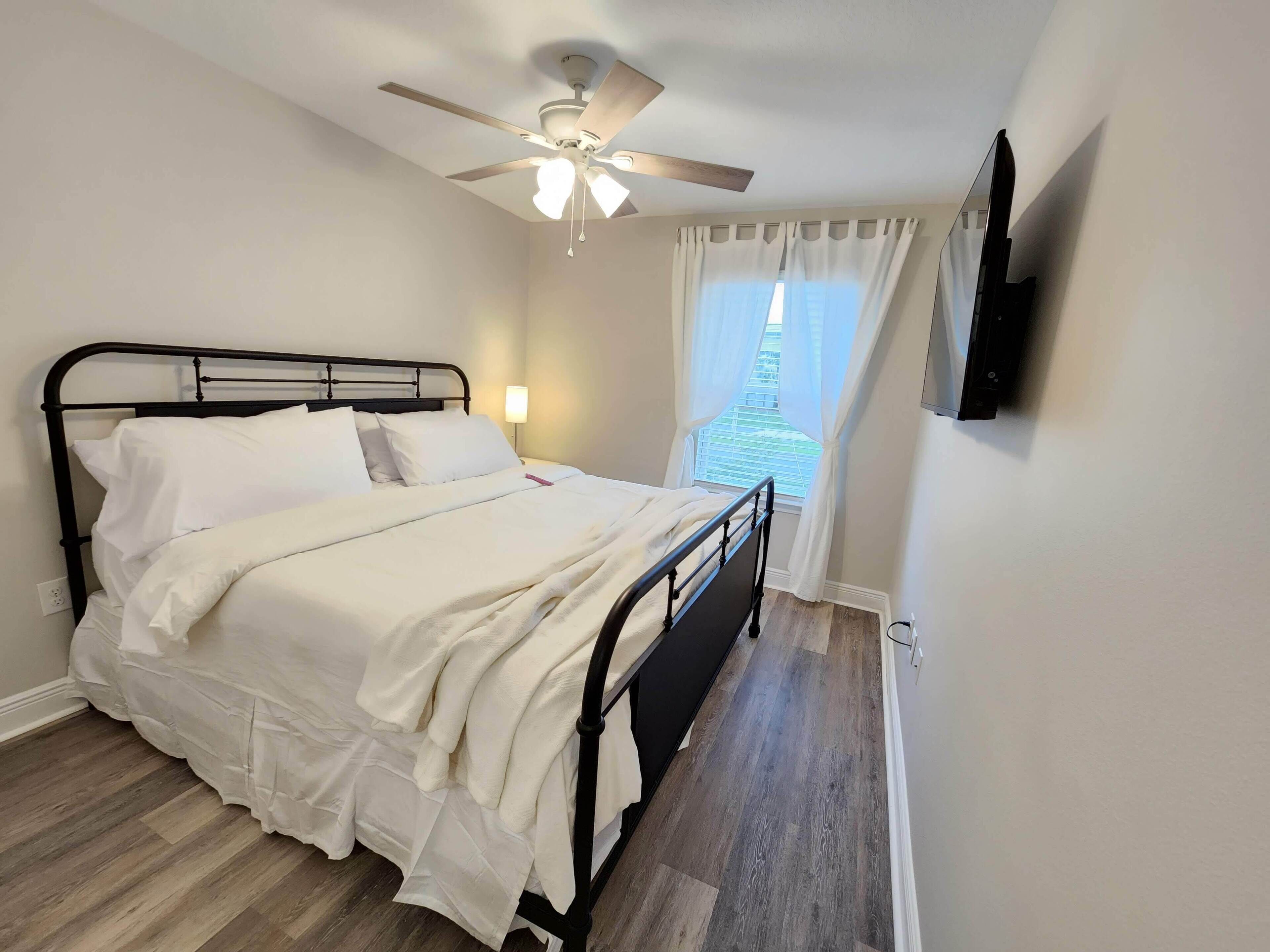 Una cama bien hecha en una habitación con ventilador de techo, una ventana con cortinas y un televisor de pantalla plana montado en la pared.