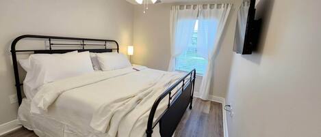 Una cama bien hecha en una habitación con ventilador de techo, una ventana con cortinas y un televisor de pantalla plana montado en la pared.