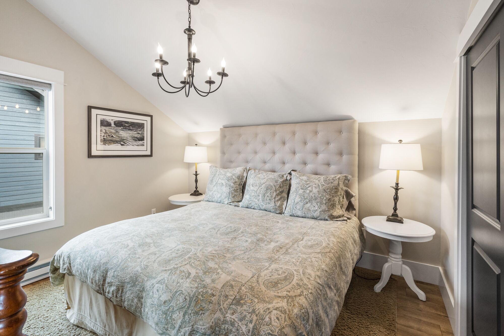 Une chambre à coucher avec une tête de lit capitonnée, un grand lit avec une literie à motifs, deux tables de chevet avec des lampes, un tableau encadré au mur et une fenêtre avec des stores.