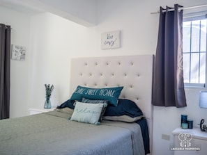 Une chambre avec une tête de lit capitonnée, un lit avec de nombreux oreillers, un vase rempli de fleurs, une pancarte indiquant « Restez bien au chaud » et une fenêtre avec des rideaux.