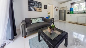 Una sala de estar moderna con un sofá gris, una mesa de centro y una zona de cocina con refrigerador y lavamanos.