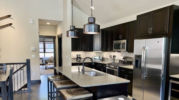 A modern kitchen with dark cabinets, stainless steel appliances, a central island with bar stools, and pendant lighting. - Luxury 3 Bedroom Mountain Townhome in Keystone (Dillon)