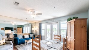 Ein Wohnzimmer mit blauem Sofa, einem Esstisch mit Glasplatte und Holzstühlen, einem Deckenventilator und einem Holzschrank.