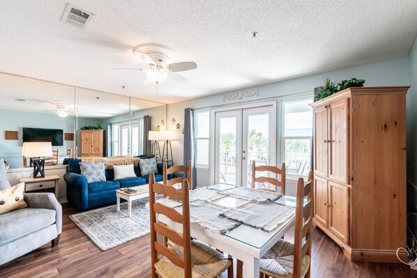 A living room with a blue sofa, a glass-top dining table with wooden chairs, a ceiling fan, and a wooden cabinet.