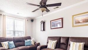Un salon avec deux canapés en cuir marron, un ventilateur de plafond, une fenêtre avec des stores et un tableau encadré accroché au mur.