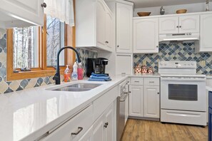 A kitchen with white cabinets, a stove, a sink, and a window with a view of trees. - Blue Skies and Cherry Pies, in Fish Creek! (Fish Creek)