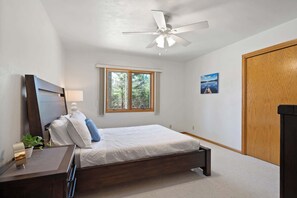 A bedroom with a bed, a ceiling fan, a window, a door, and a small bedside table with a lamp.