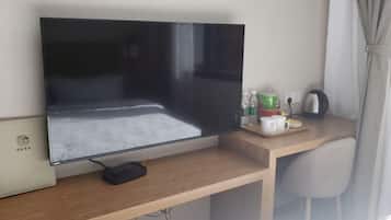 A flat-screen TV mounted on a wooden desk with a power strip, a white chair, a small table with cups and a kettle, and a window with curtains.