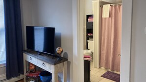 Ein Zimmer mit einem Fernseher auf einem Holzboden, einem Globus und einer Scrabble-Box. Ein Badezimmer ist sichtbar, mit einem rosa Duschvorhang und einer Toilette.
