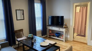 Ein Wohnzimmer mit Holztisch, einer Couch, einem Fernseher auf einem Schrank und einer Tür zu einem Badezimmer.