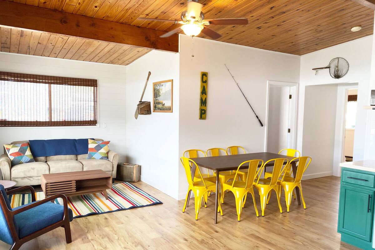 A cozy living room with a wooden ceiling, a sofa with colorful cushions, a striped rug, and a dining area with yellow chairs and a wooden table.