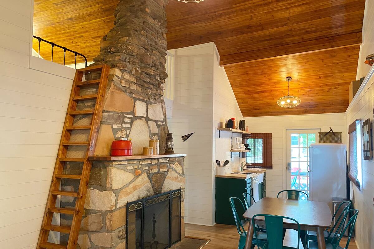 A kitchen with a stone fireplace, wooden ceiling, and a dining area with green chairs.