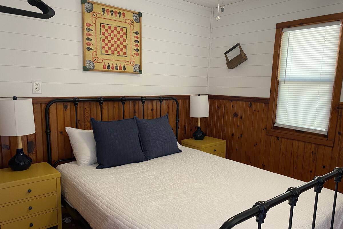 A neatly arranged bedroom with a bed, two bedside tables, a lamp, a wall clock, and a window with blinds.