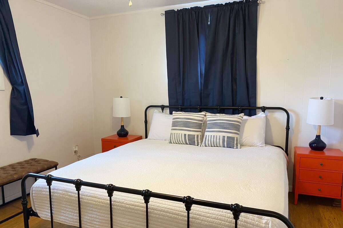 A neatly made bed with a white duvet, two bedside tables with lamps, a bench, and a window with dark curtains.