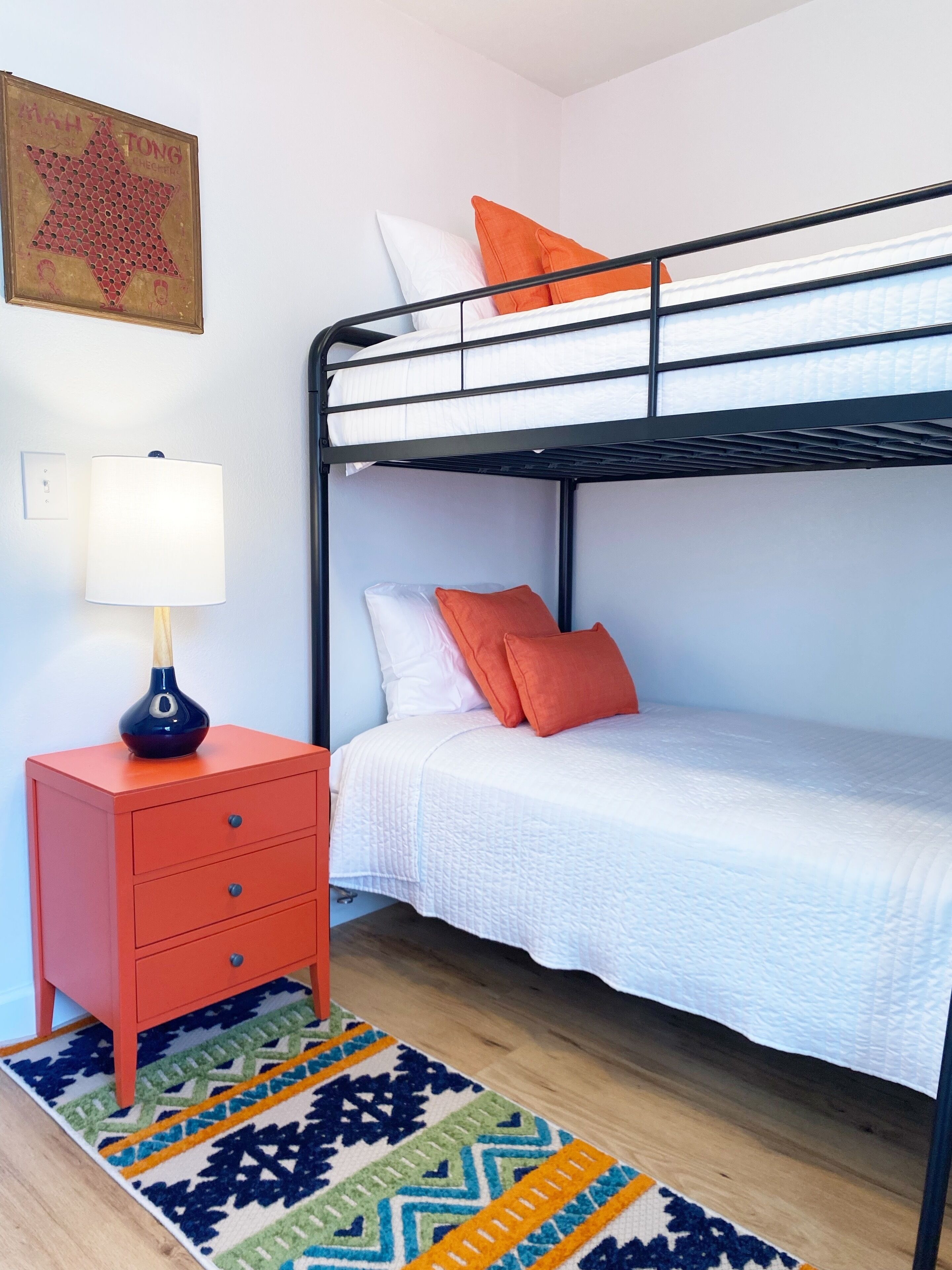 Un lit superposé avec une literie blanche et des oreillers orange, une commode rouge avec une lampe, et un tapis à motifs sur un plancher en bois.
