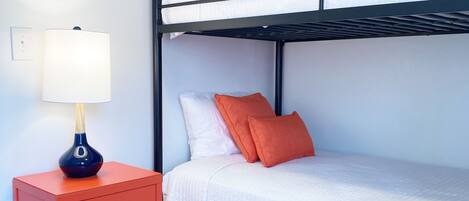 Un lit superposé avec une literie blanche et des oreillers orange, une commode rouge avec une lampe, et un tapis à motifs sur un plancher en bois.