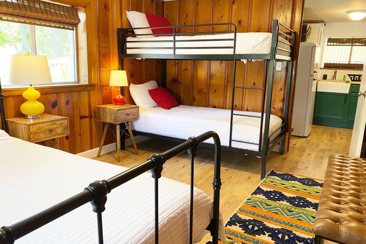 A bunk bed room with a wooden wall, a bedside table with a lamp, and a colorful rug.