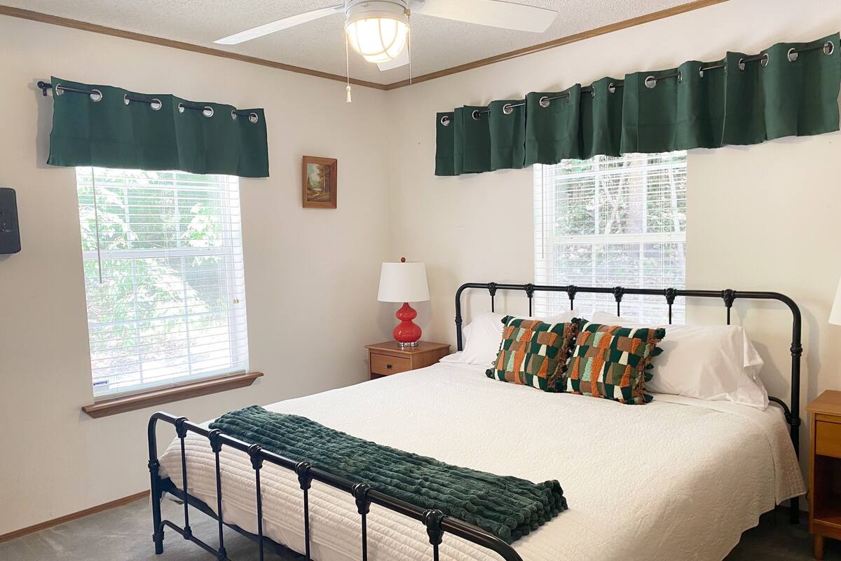A neatly arranged bedroom with a bed, two bedside tables, a nightstand, a ceiling fan, and two windows with curtains.