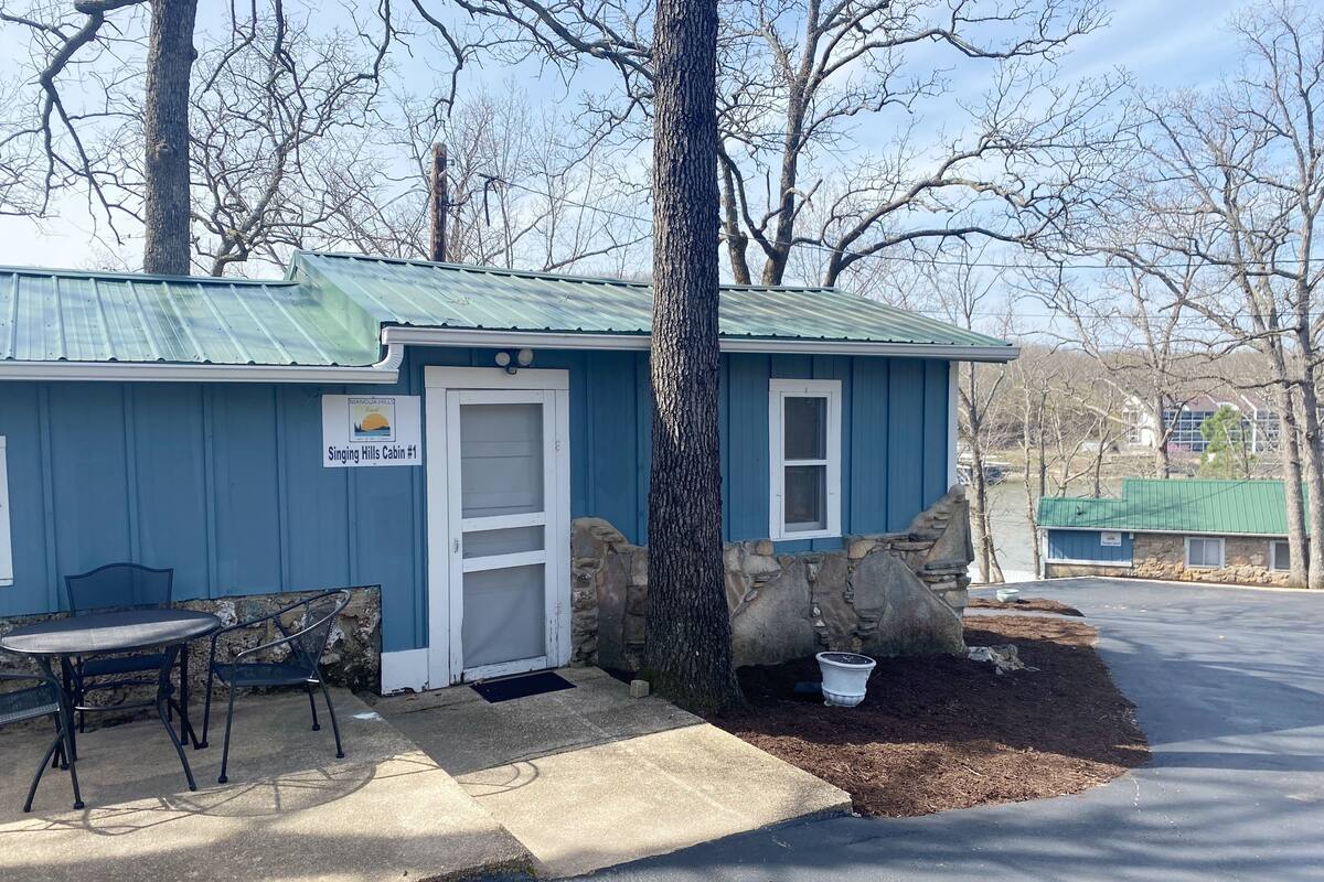 A small cabin with a blue exterior, a white door, and a stone wall. There is a table and chairs outside.