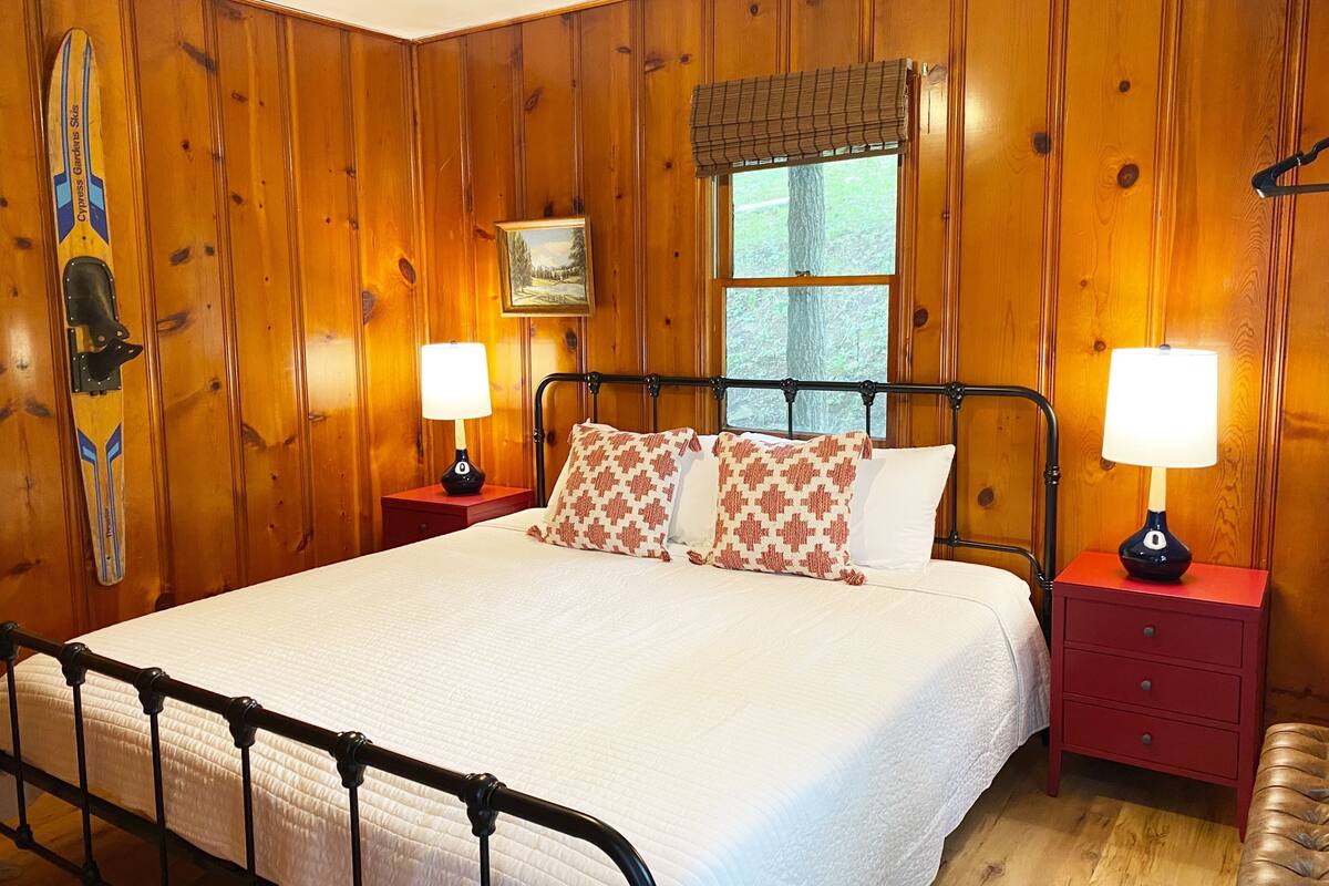 A wooden-paneled bedroom with a bed, two bedside tables with lamps, a window with a view, and a framed picture on the wall.