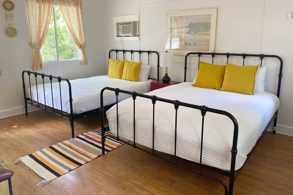 A room with two beds, a window with curtains, a wall-mounted air conditioner, and a framed picture.