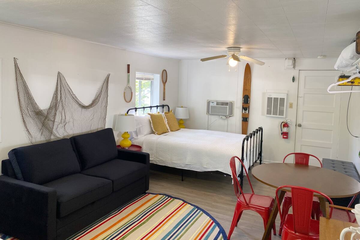 A compact room with a bed, a sofa, a dining table with chairs, a ceiling fan, and a surfboard on the wall.