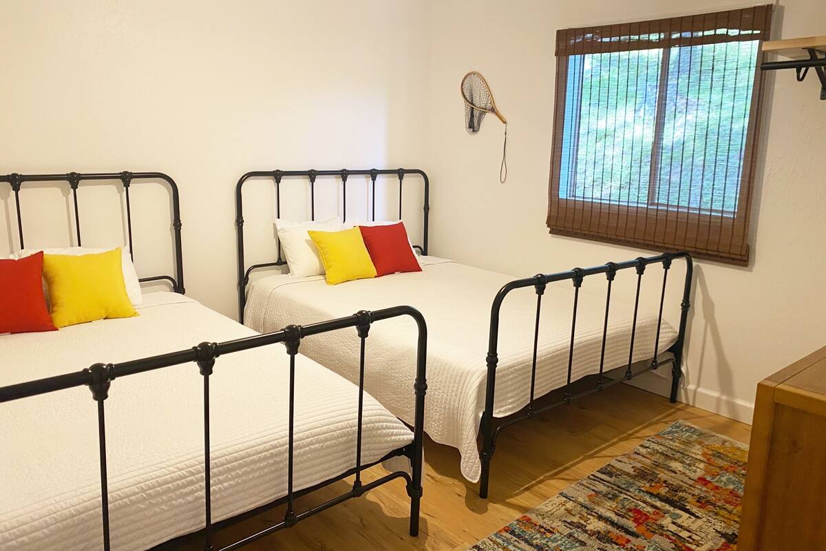 A room with two single beds, a window with blinds, a fan, and a patterned rug.