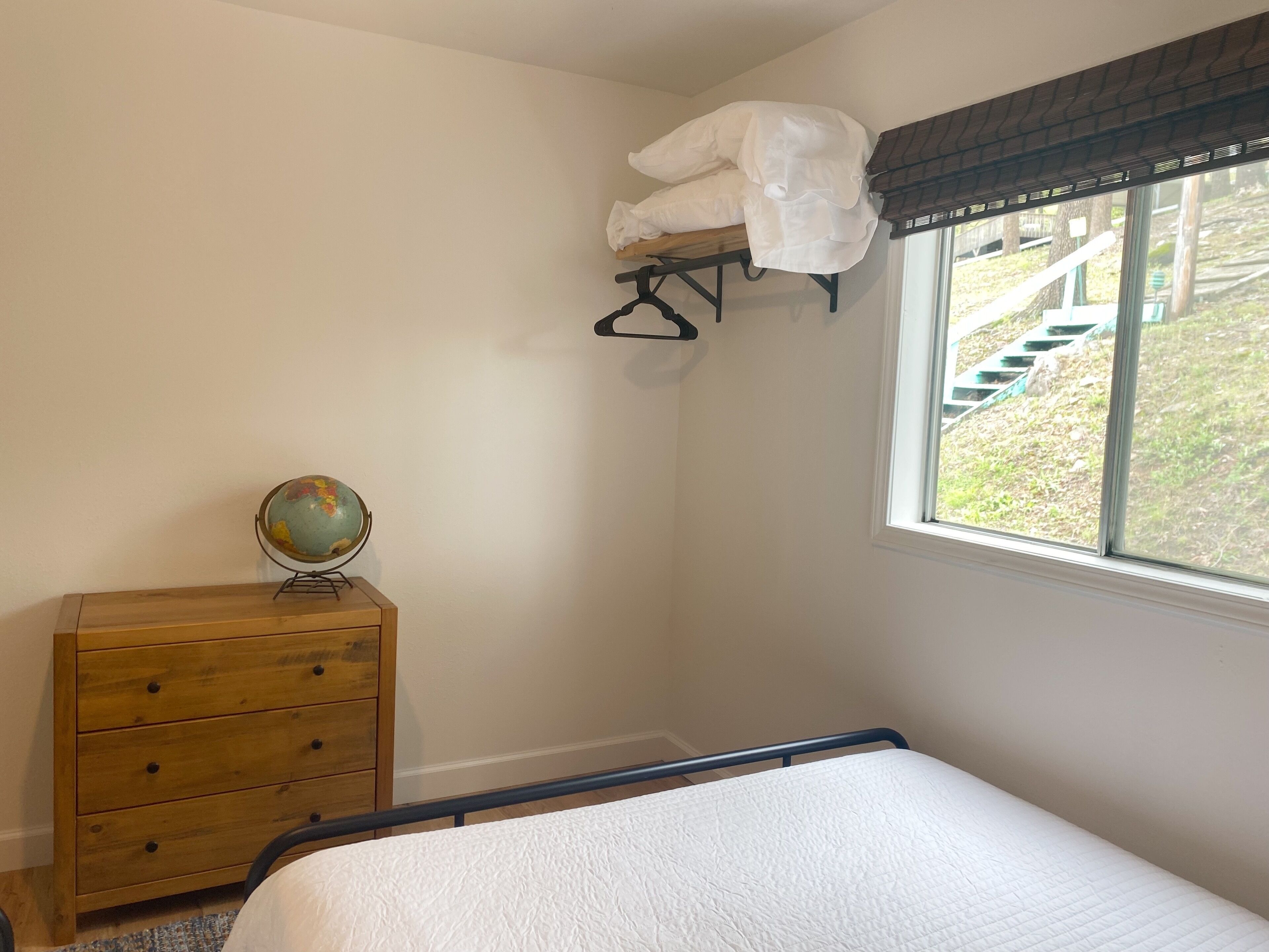 A bedroom with a bed, a wooden dresser, a globe, and a window with a view of a staircase.