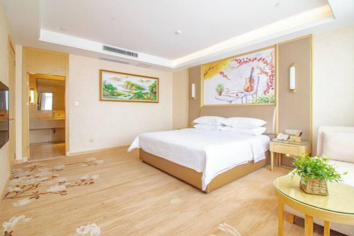 A hotel room with a bed, a bedside table, a chair, and a small table with a plant. There is a painting on the wall and a door leading to another room.