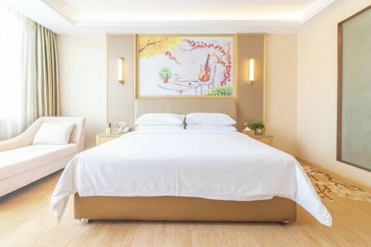 A hotel room with a large bed, a sofa, a painting on the wall, and a large window.
