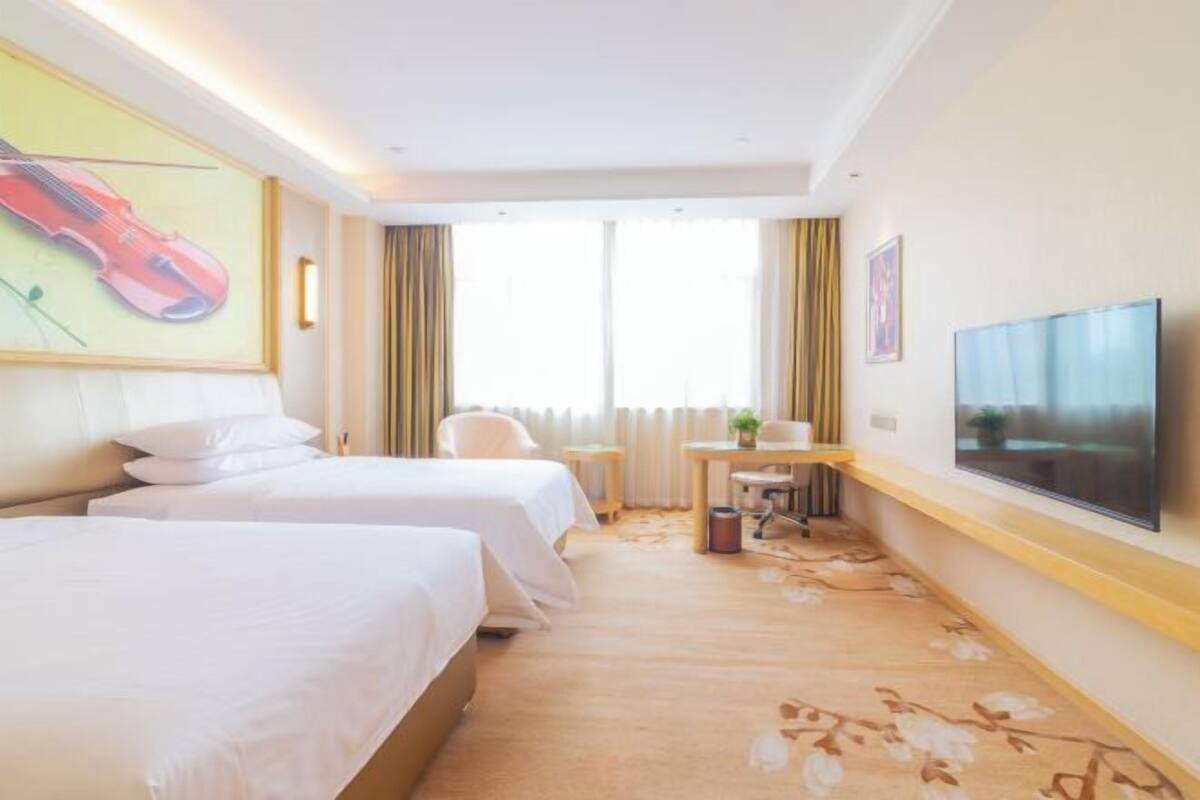 A hotel room with two beds, a desk, a chair, a TV, and a large window with curtains.