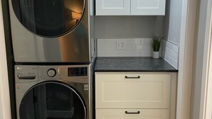 Une cuisine compacte équipée d’un lave-linge et d’un sèche-linge à double tambour en acier inoxydable, de meubles blancs et d’un petit plan de travail agrémenté d’une plante en pot.