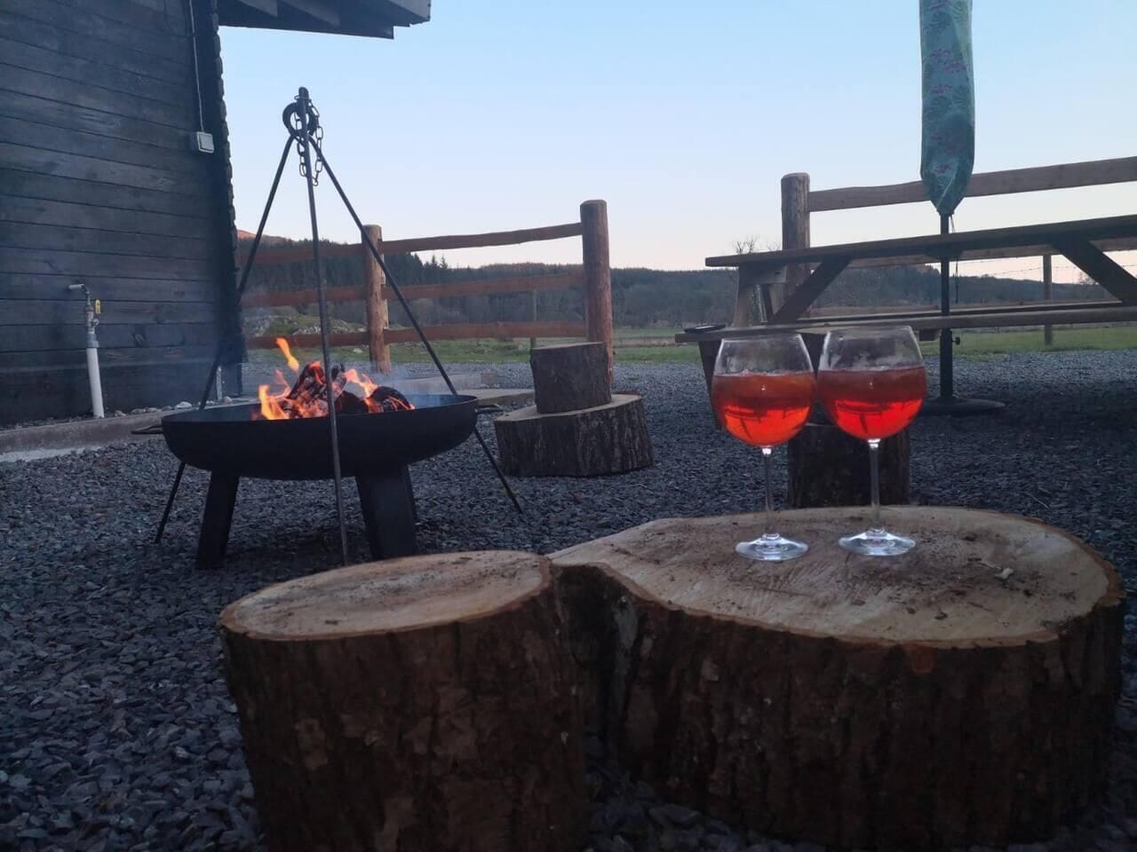 Ambientazione esterna rustica con un braciere e due bicchieri di vino rosso poggiati su tronchi di legno.