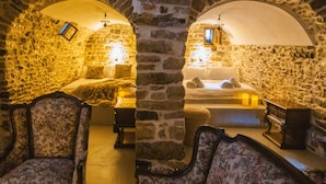 A stone-walled room with a bed, two chairs, a small table, and a hanging light fixture.
