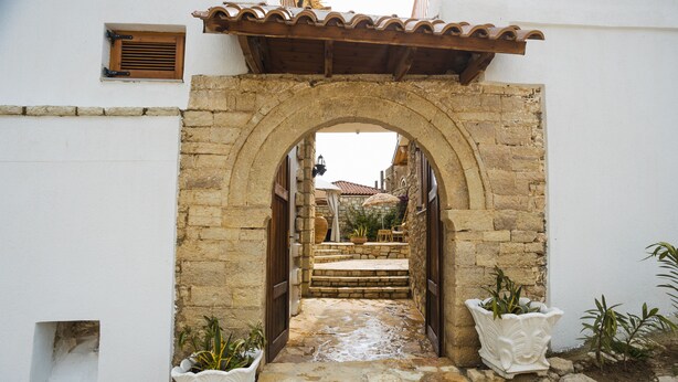 An arched stone doorway leading to a courtyard with steps, potted plants, and a tiled roof in the background.
