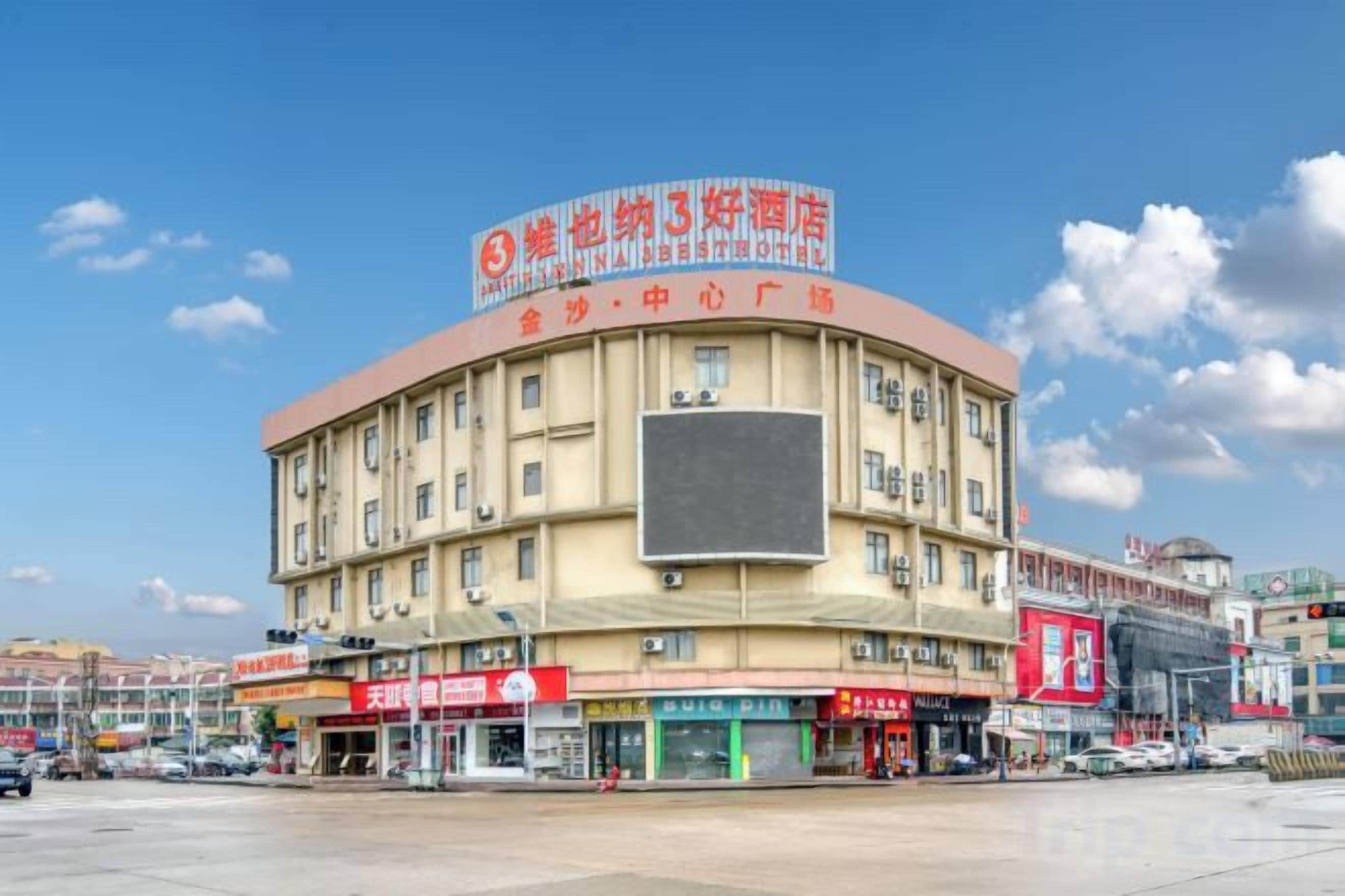 Un hotel de varios pisos con un gran letrero central, varias tiendas a nivel de la calle y un cielo despejado.