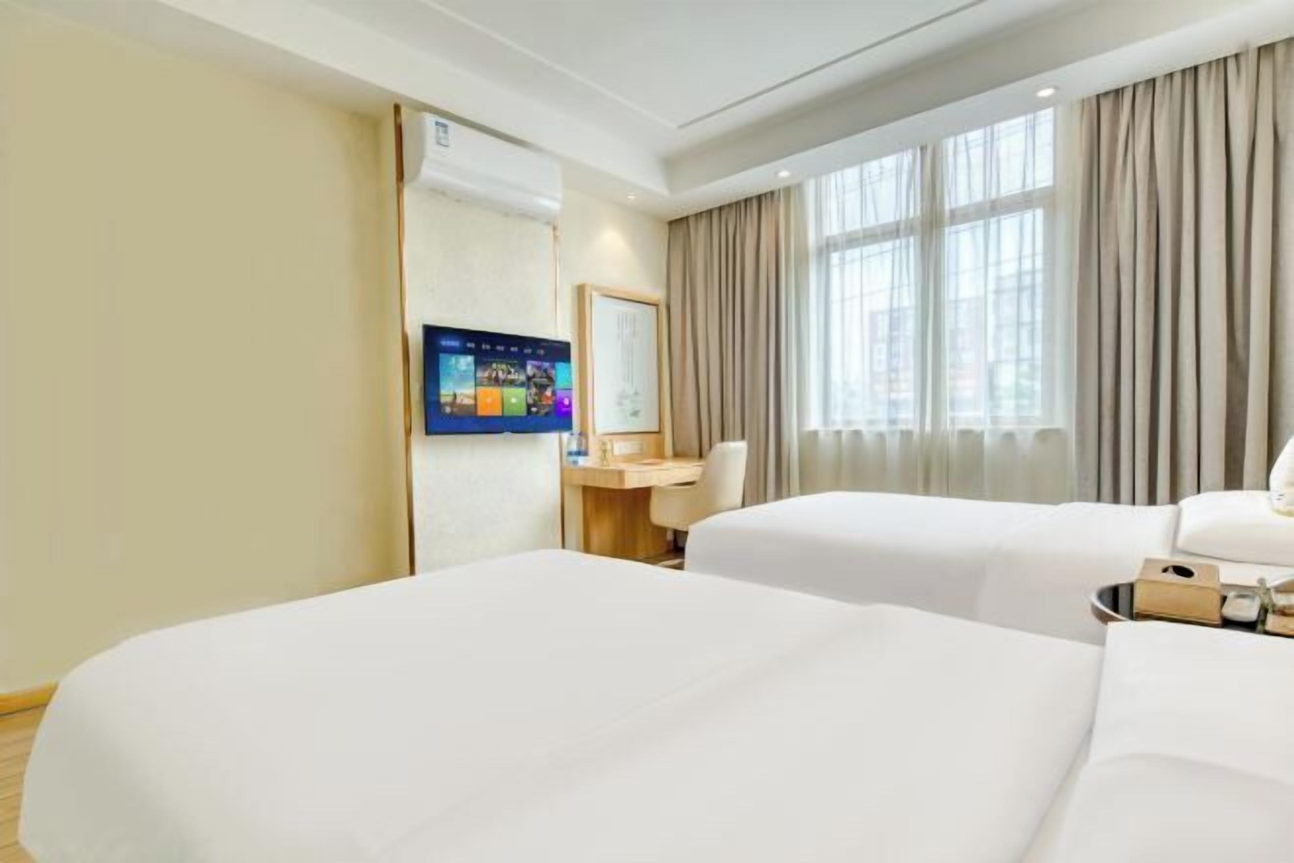 A hotel room with two beds, a desk, a TV, and large windows with curtains.