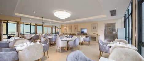A spacious dining area with round tables and modern chairs, large windows offering a view, and a central chandelier.