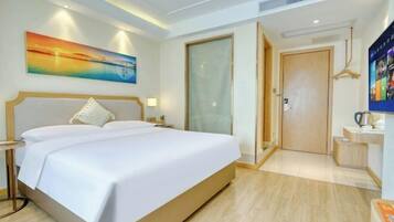 A hotel room with a large bed, a painting on the wall, a desk with a kettle, and a door leading to another room.