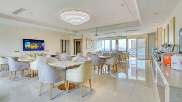 A spacious dining area with multiple tables and chairs, a television screen, and a large chandelier.