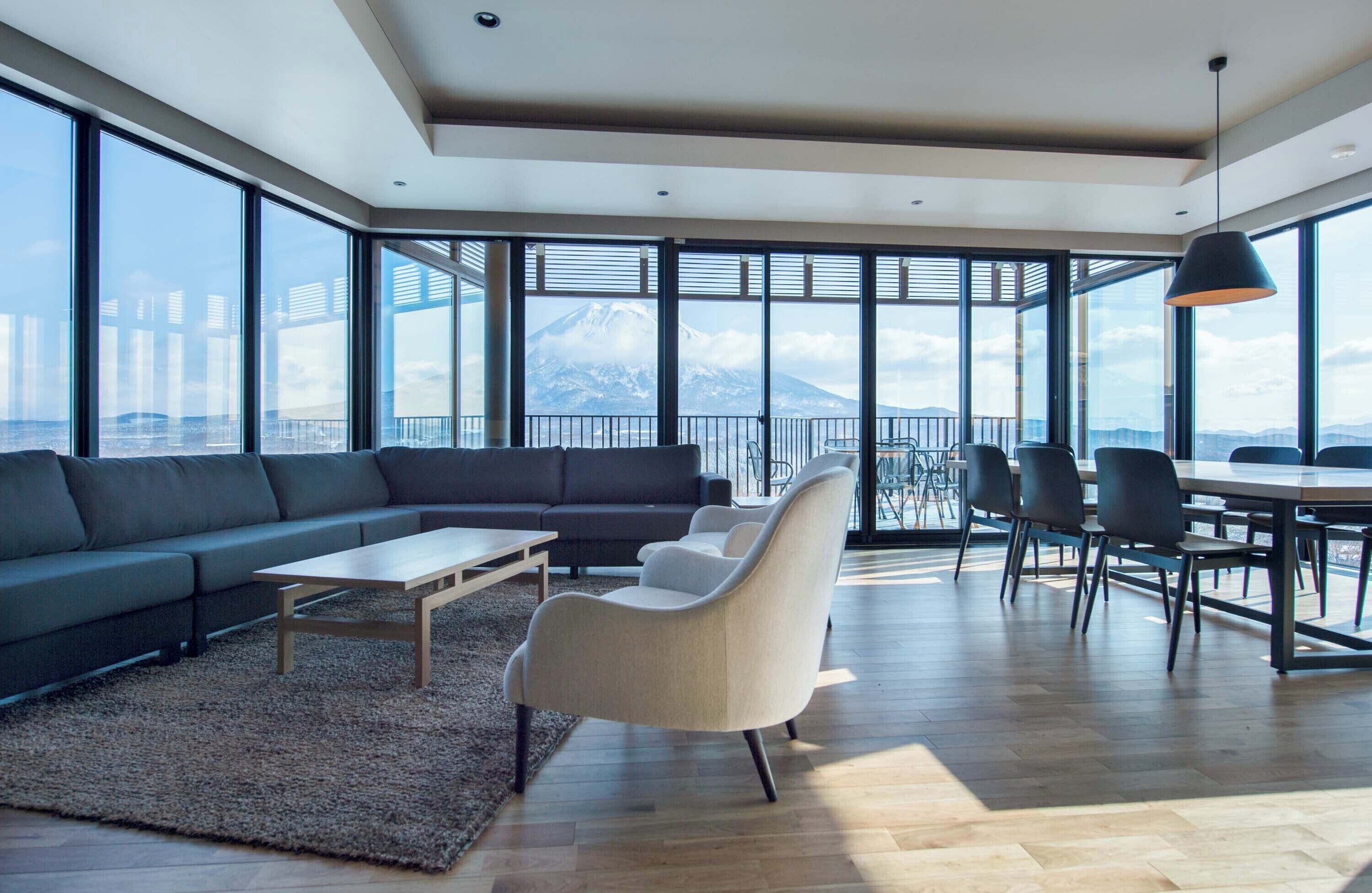 A modern living room with a sofa, chairs, a coffee table, and a dining area with chairs and a table. Large windows provide a view of a mountain.