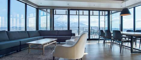A modern living room with a sofa, chairs, a coffee table, and a dining area with chairs and a table. Large windows provide a view of a mountain.