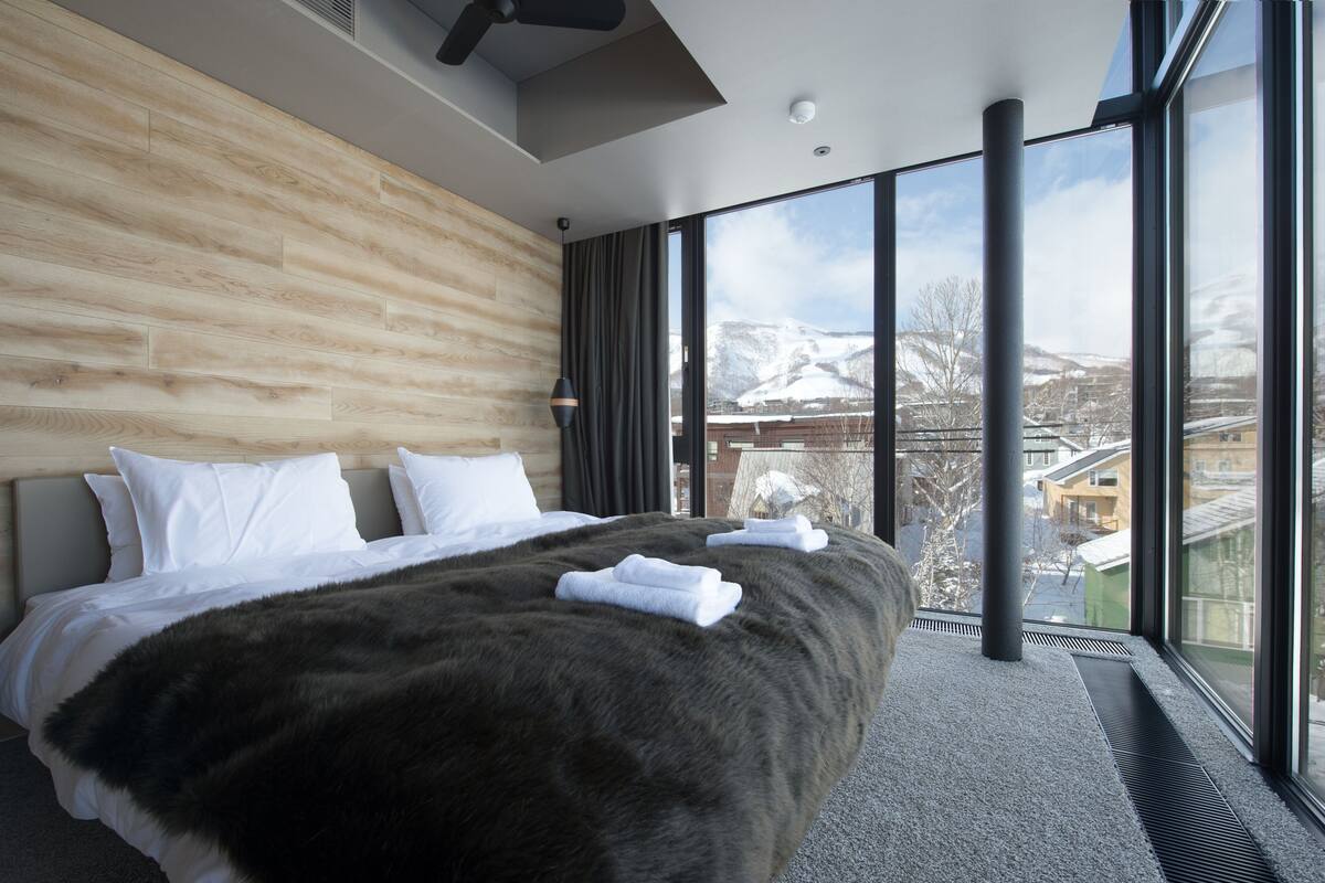 Una habitación de hotel moderna con una cama grande, cabecero de madera y vistas a un paisaje nevado a través de amplios ventanales.