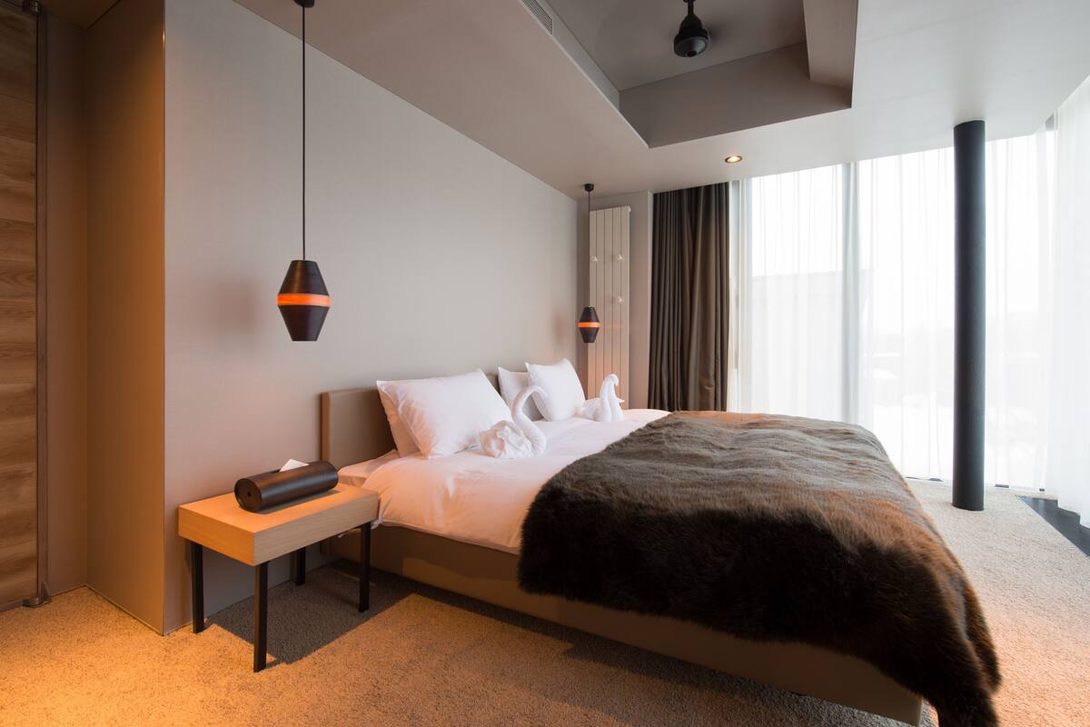 Habitación de hotel moderna con una cama grande, una mesita de noche de madera, una lámpara negra y vistas a la ciudad a través de amplias ventanas.