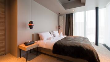 A modern hotel room with a large bed, a wooden nightstand, a black lamp, and a view of the city through large windows.