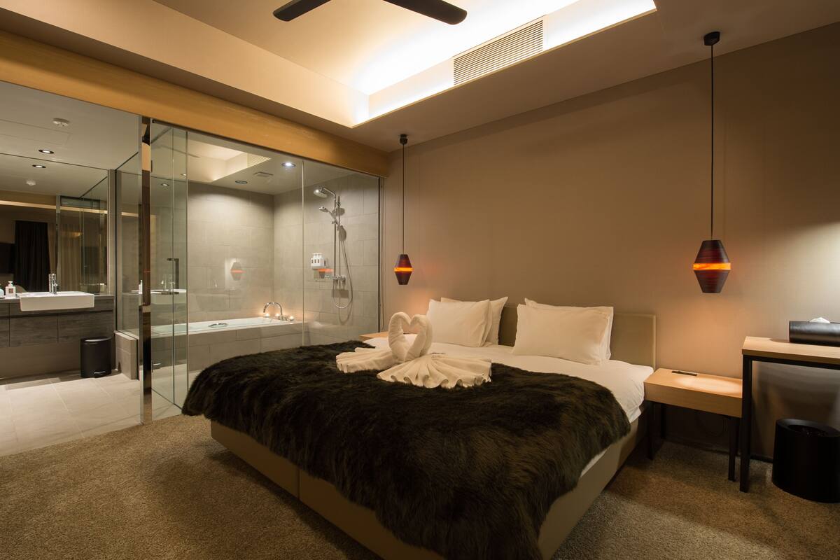 Una habitación de hotel moderna con una cama grande, zona de ducha con cerramiento de vidrio, un escritorio y una silla.