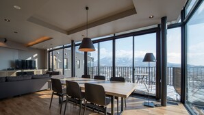 A modern kitchen with a large island, a dining area with a wooden table and chairs, and large windows offering a view of a snowy mountain. - Aspect Luxury Apartments by H2 Life (Kutchan)